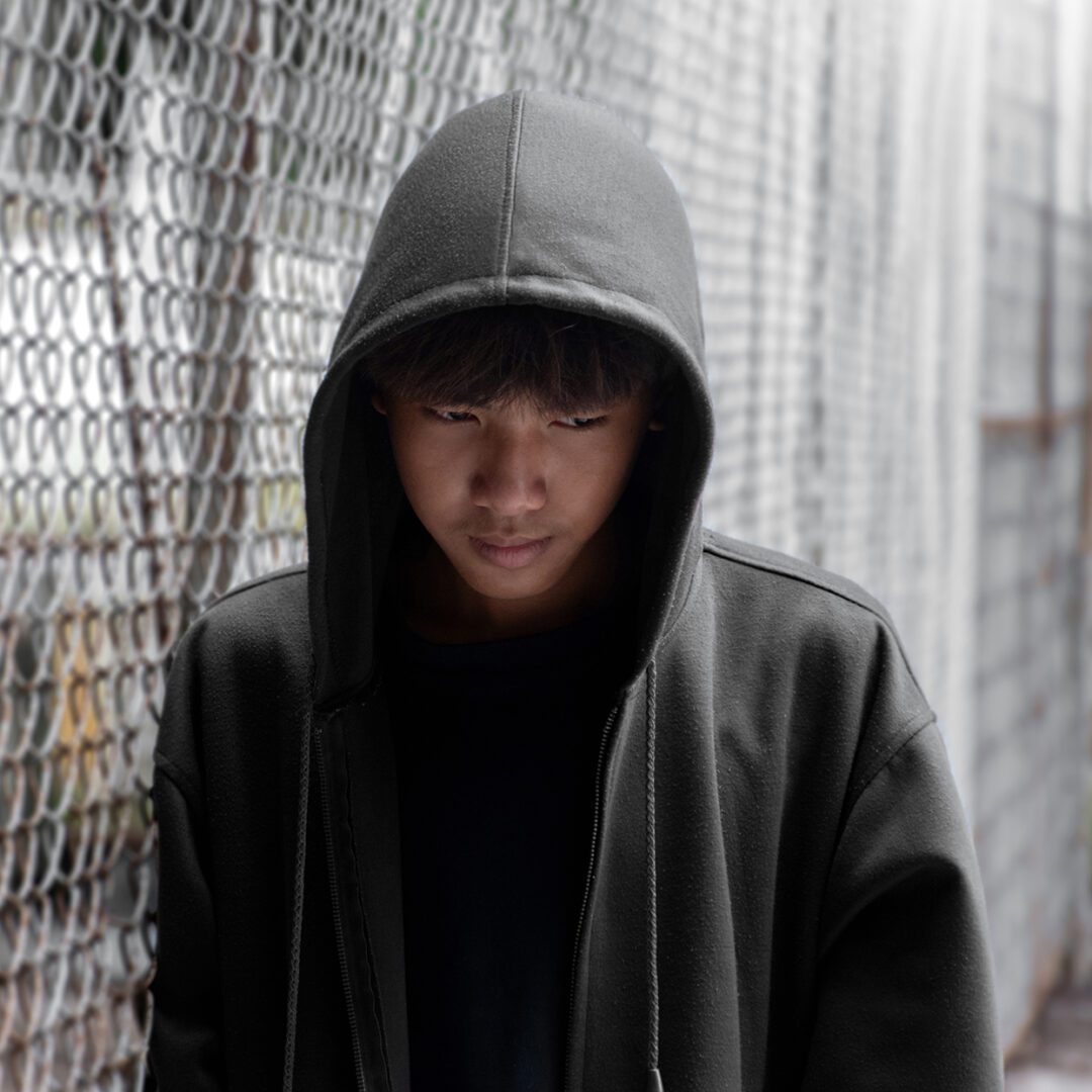 Person in dark hoodie standing near a chain-link fence, looking serious.