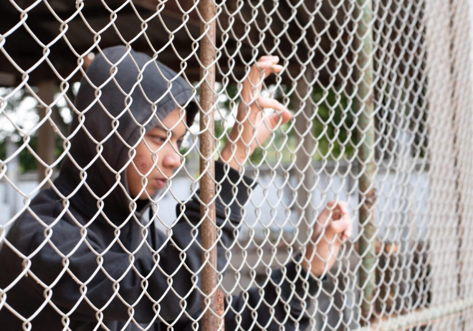 Woman in hijab gripping chain-link fence with a somber expression.